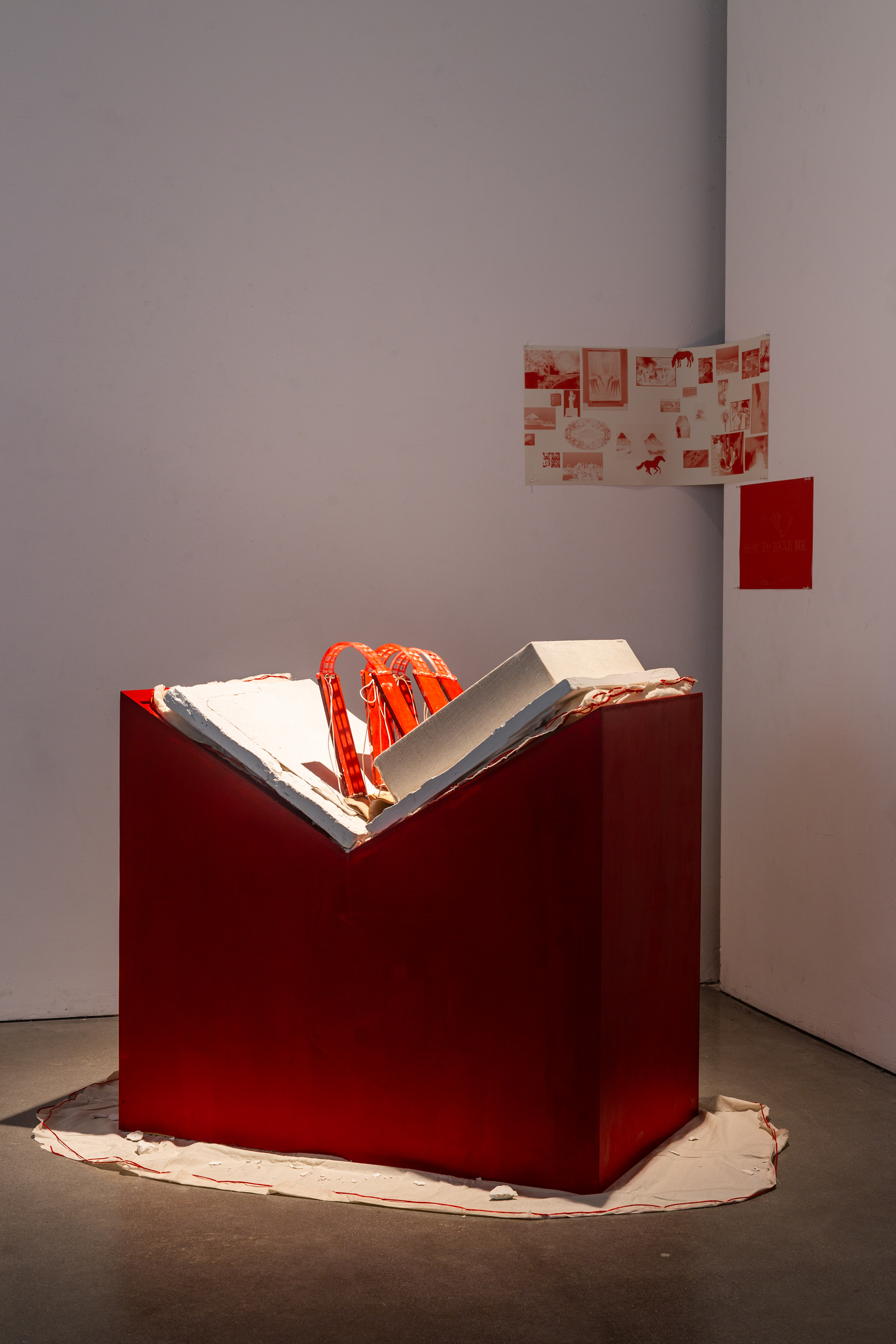 A red geometric sculpture with white and gray slabs forming a book protruding from the top, displayed against a plain white wall.